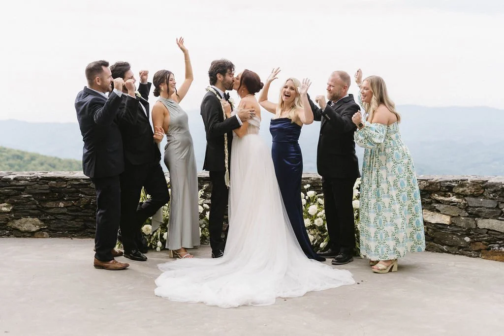 Bride and groom hug while family and friends cheer around them on a stone wall overlook.