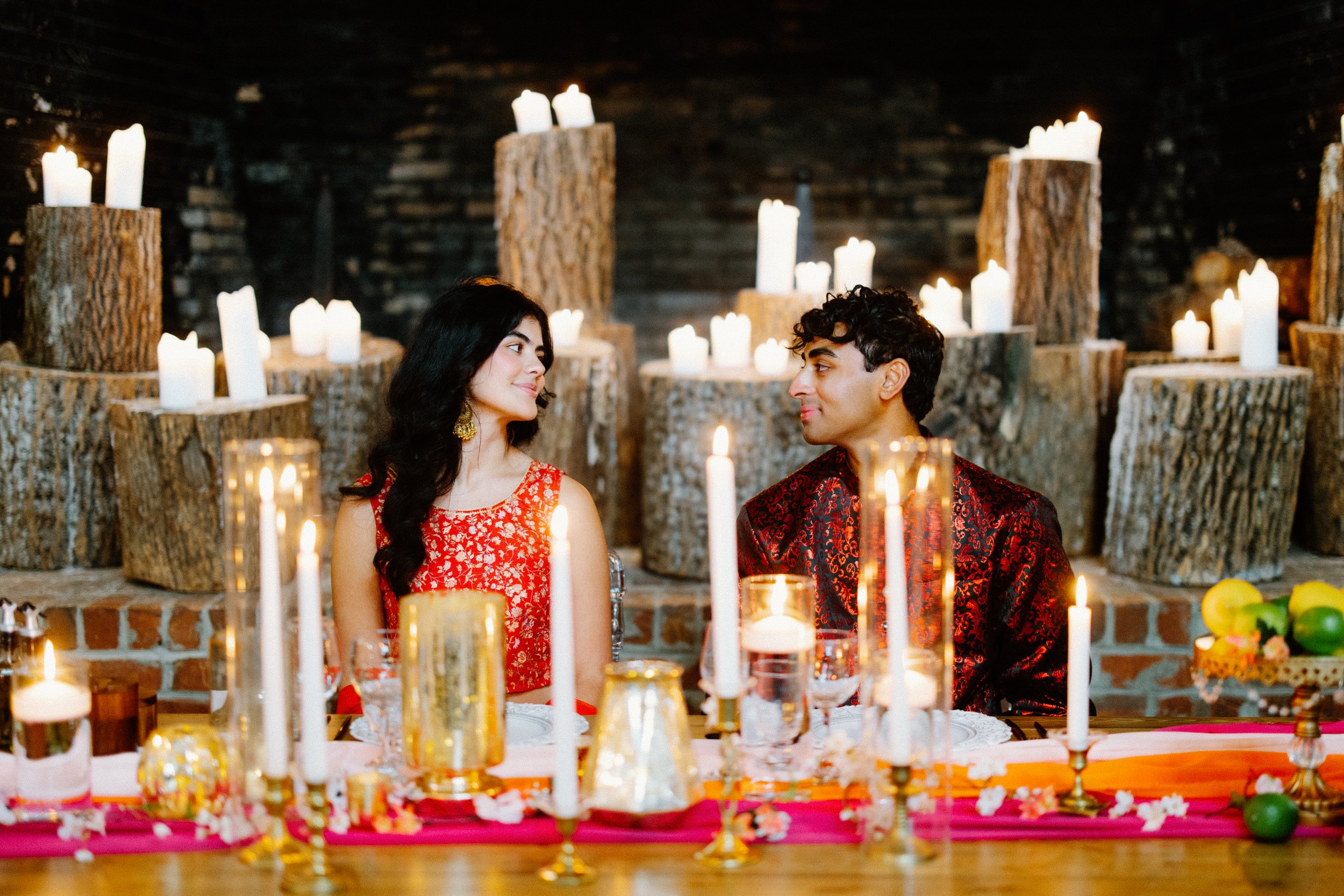 Couple sits at a long table, gazing at each other amid tall candles and rustic wood candle stands in the background.