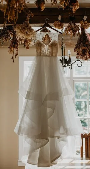 Wedding dress hanging in front of a window with dried flowers overhead