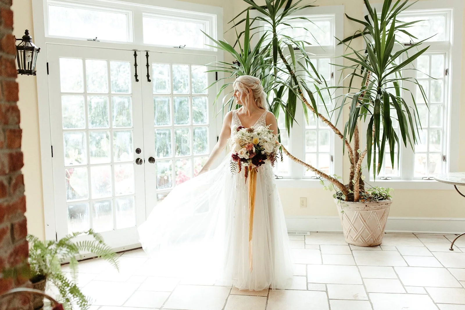 Bride standing indoors in her wedding dress while holding a bouquet beside bright windows and a palm plant.