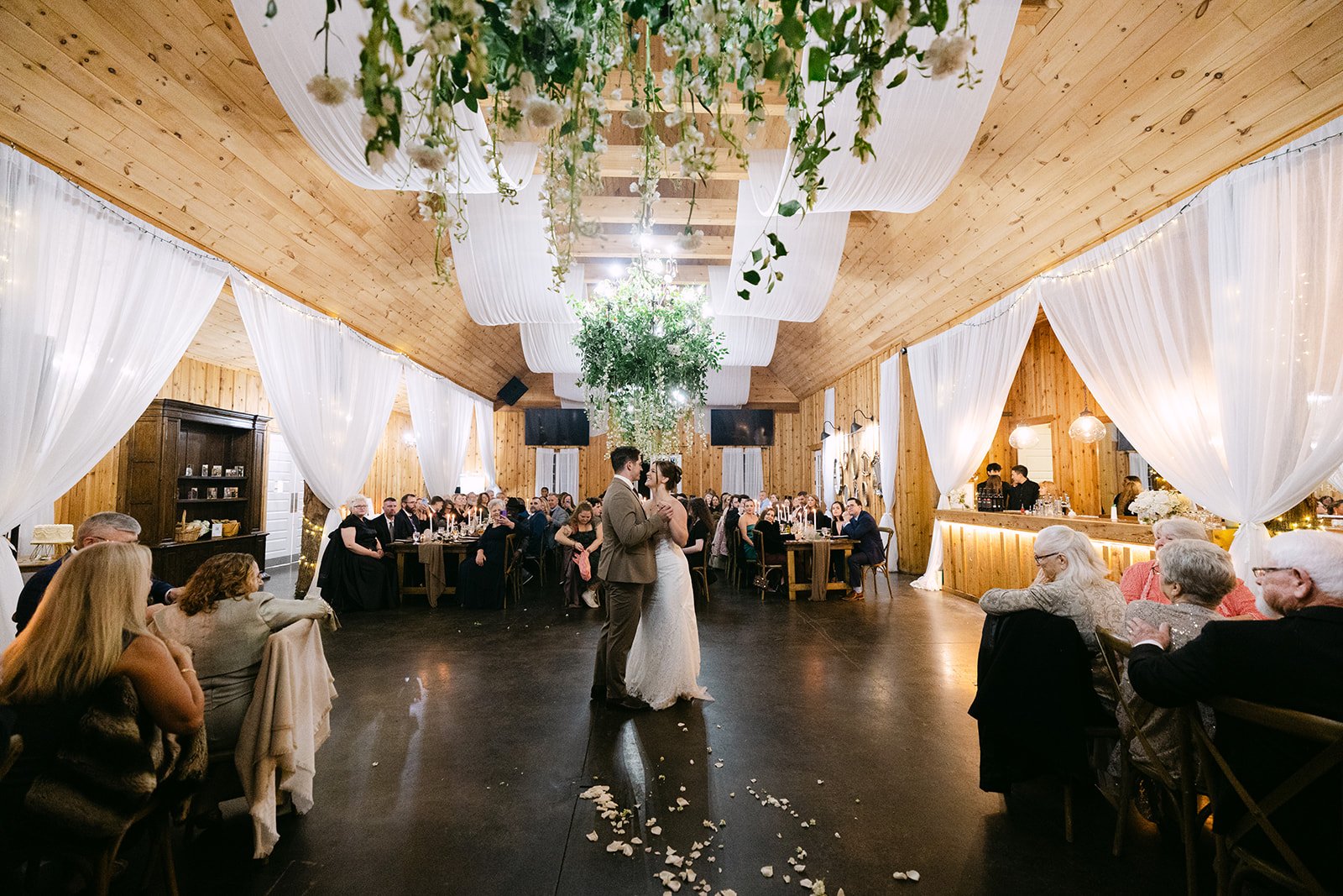 Bride and groom sharing their first dance inside a wooden reception hall with white draping and hanging greenery.