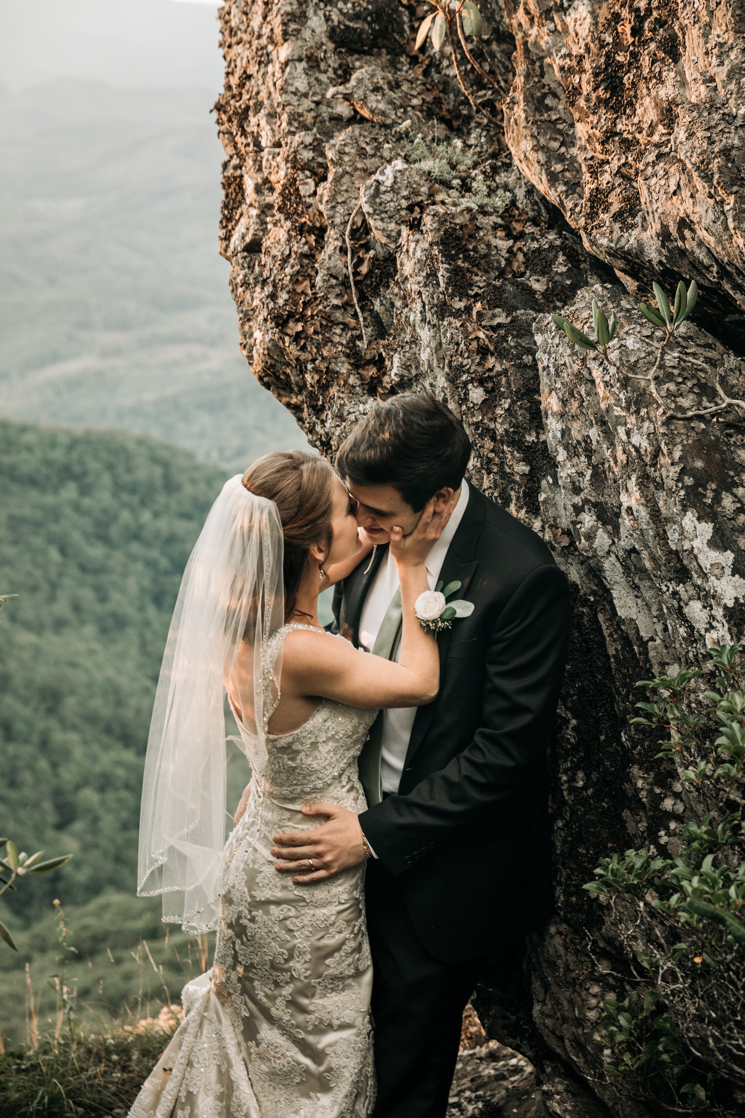 Bride and groom kissing beside a rocky mountain cliff during a romantic overlook wedding portrait.