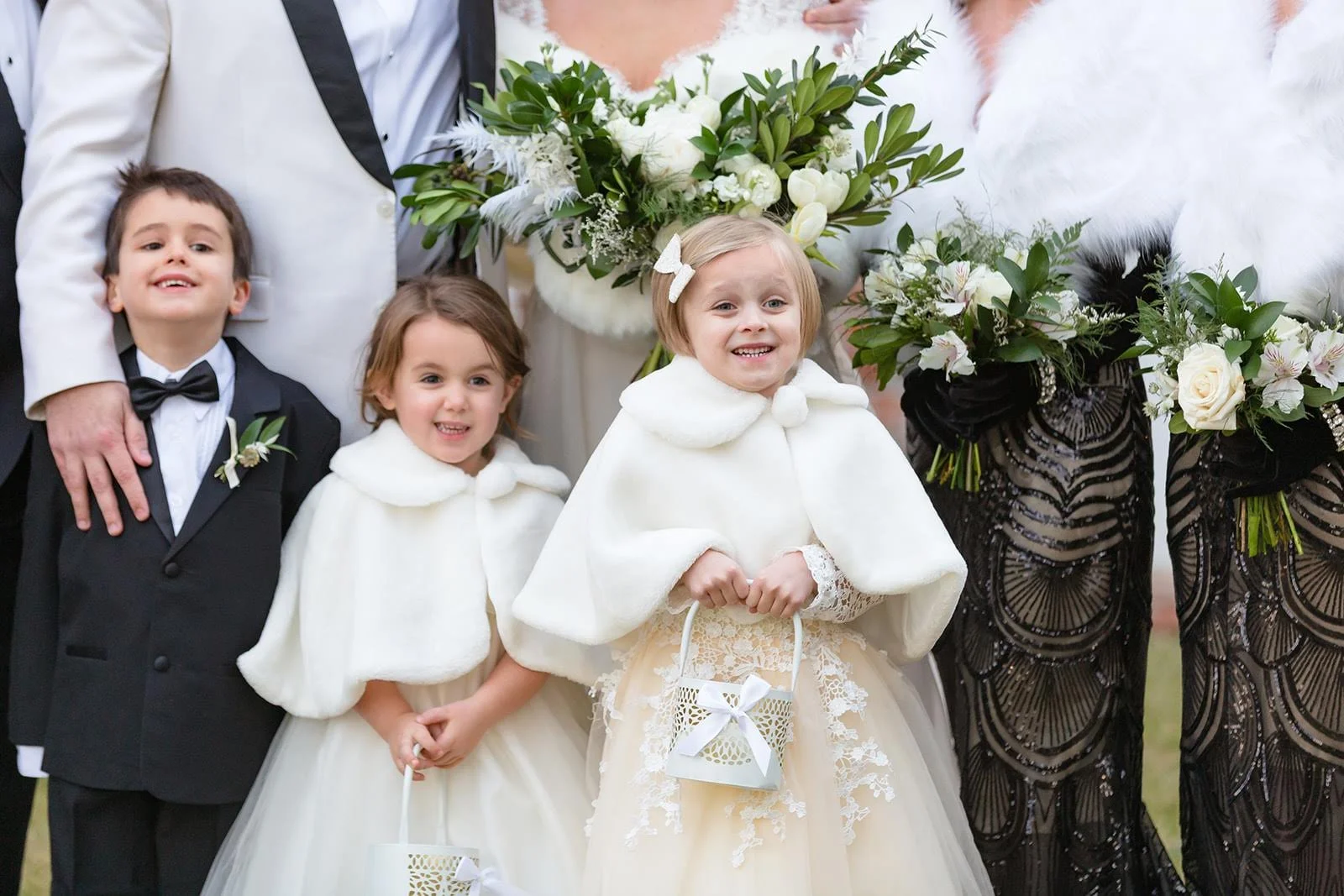 Two flower girls wearing white capes standing in front of the wedding party holding small baskets.