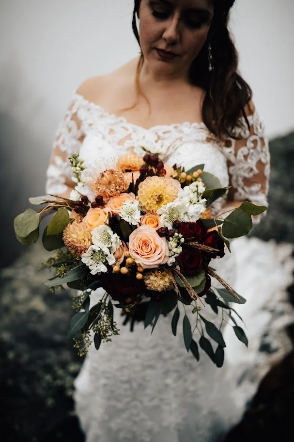Bride in a lace wedding dress holding a fall bouquet with orange and burgundy flowers and greenery.