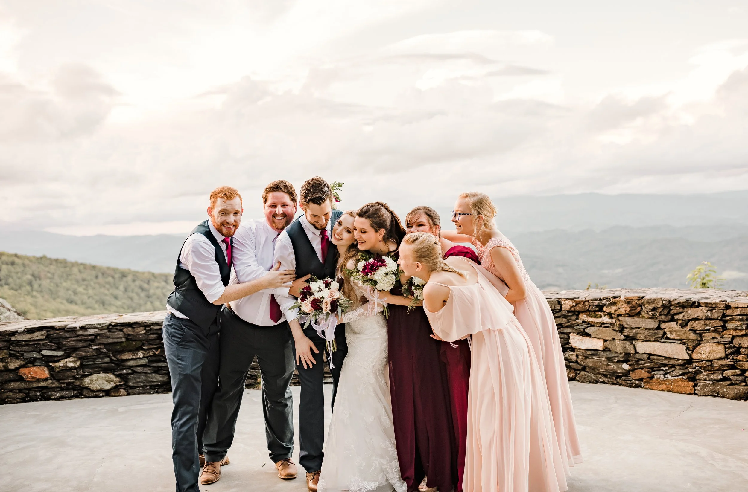 A happy wedding party poses for a group hug. The bride and groom are embraced by three groomsmen and four bridesmaids, all smiling warmly.