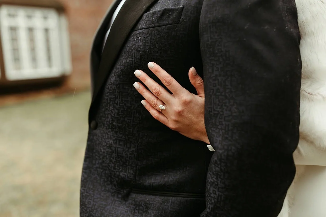 Closeup of the bride’s hand showing a wedding ring resting on the groom’s black suit jacket.