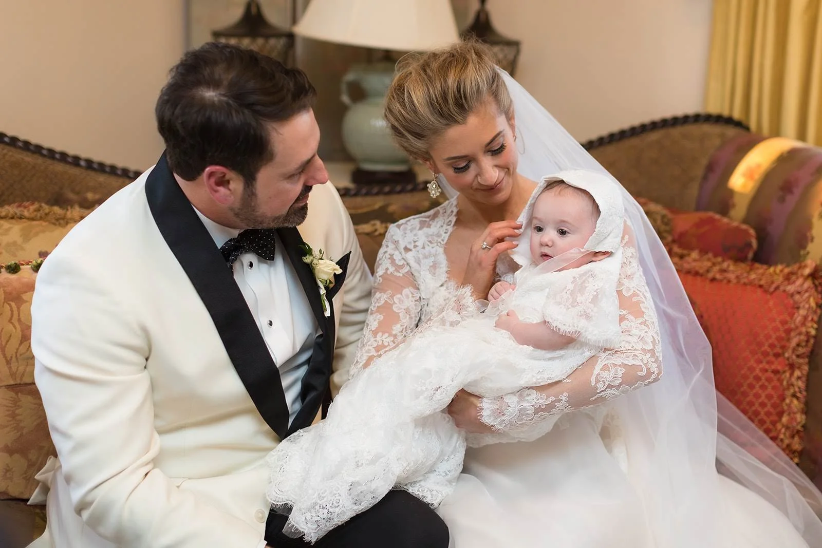 Bride and groom holding a baby in a christening gown during an indoor wedding family portrait.