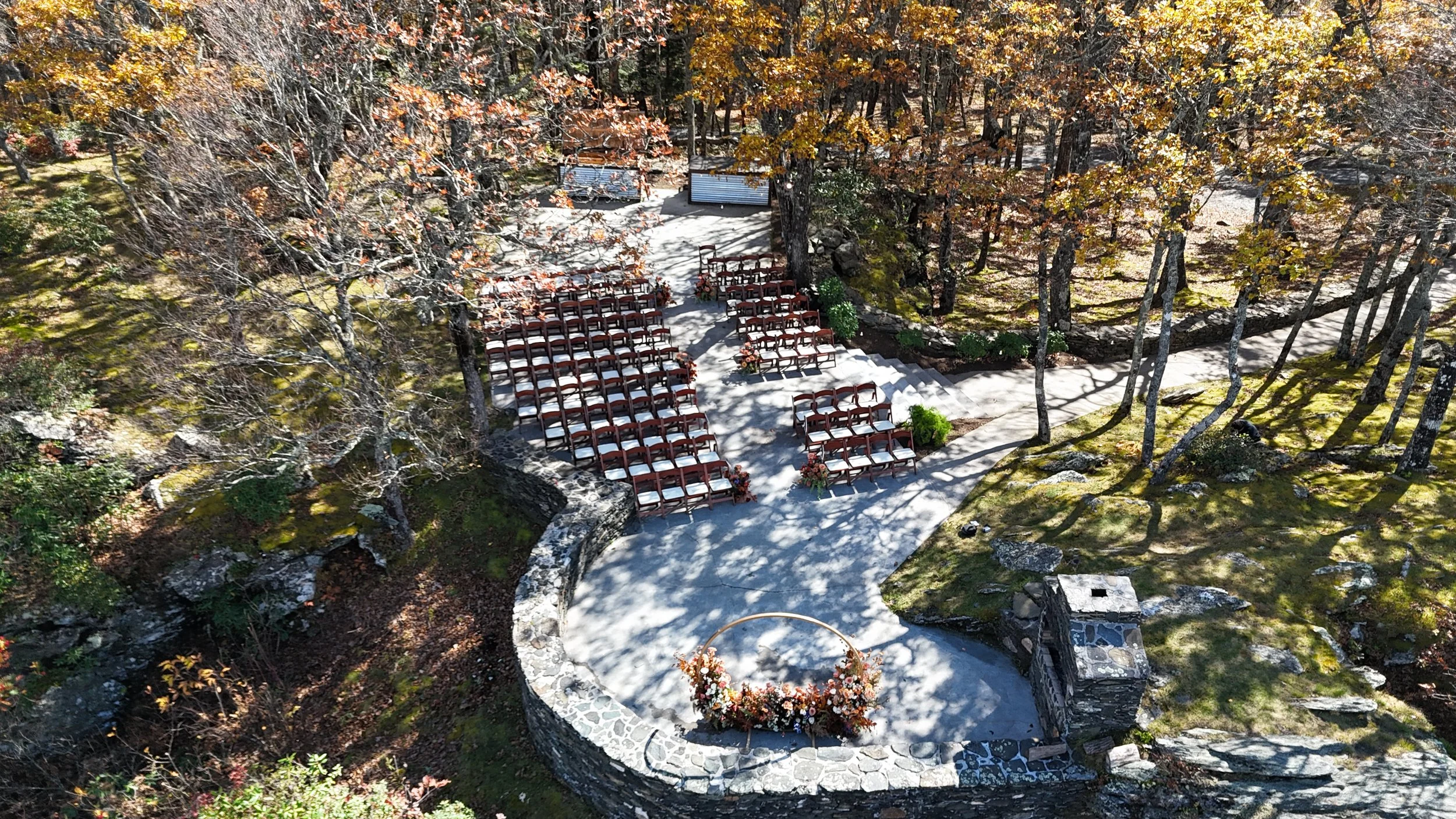 Aerial view of an outdoor ceremony site with rows of wooden chairs on a curved stone terrace among fall trees.