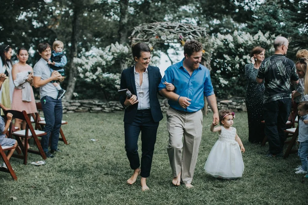 A smiling couple walks down a grassy aisle after their wedding ceremony, arm in arm, followed by a young flower girl. Wedding guests are seated on either side, observing the processional in an outdoor setting.