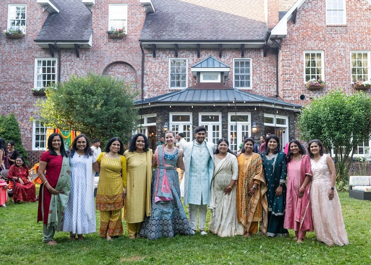 Group of guests in colorful outfits pose on the lawn in front of a brick estate, smiling toward the camera.