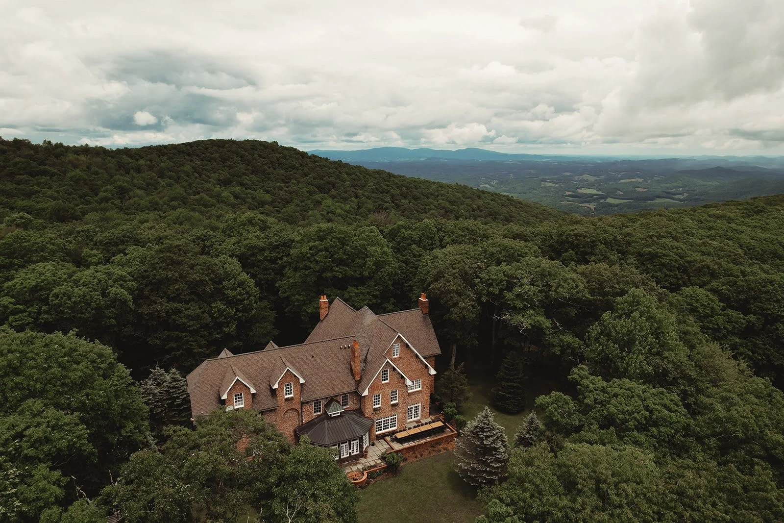 Aerial view of a brick estate surrounded by dense green forest with layered mountain views in the distance