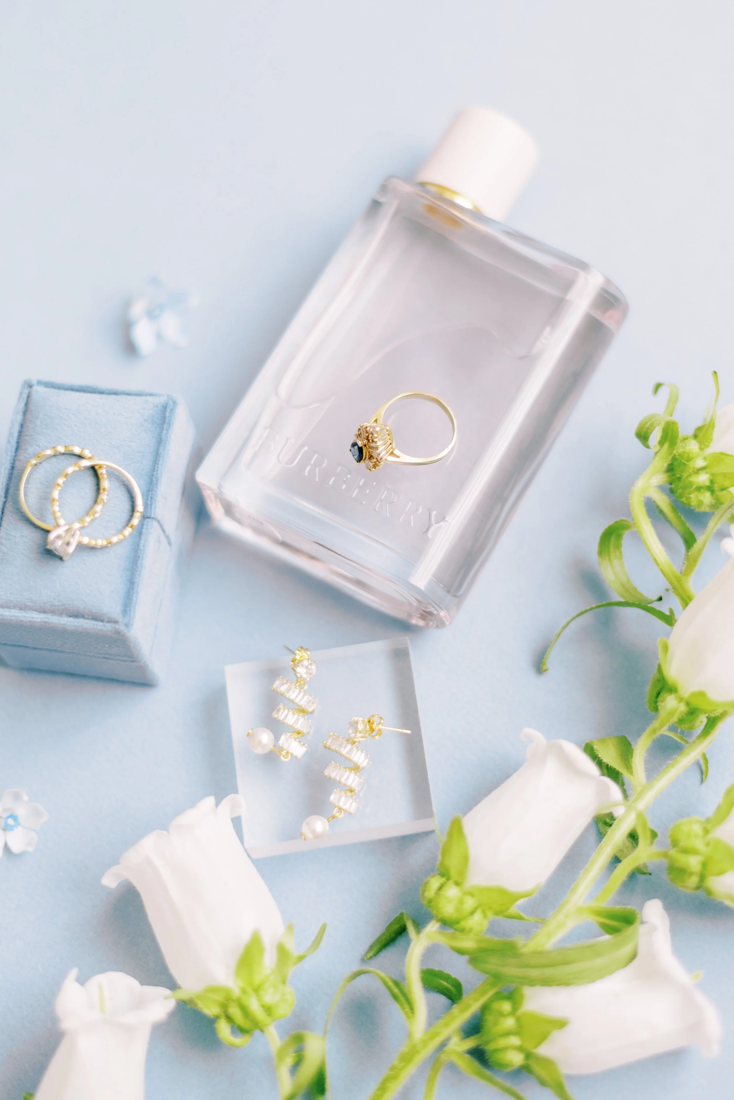 Engagement ring resting on a clear perfume bottle beside a blue ring box on a light background.