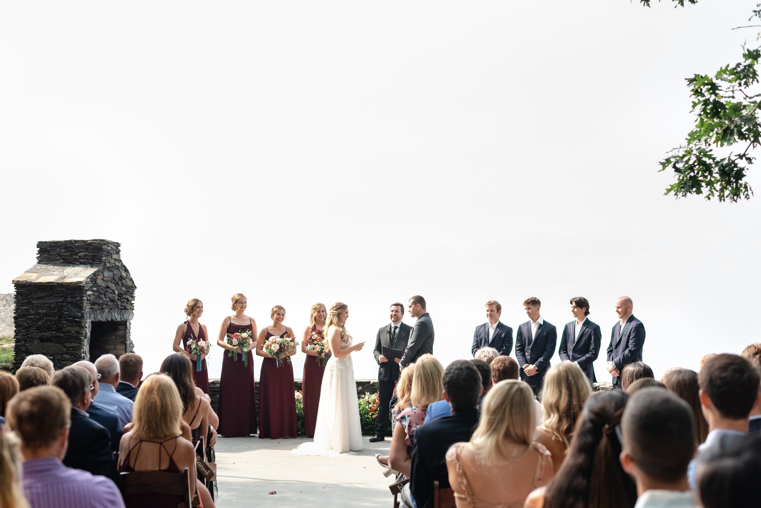 Outdoor mountain wedding ceremony with bride, groom, wedding party, officiant, and seated guests overlooking a bright scenic backdrop.