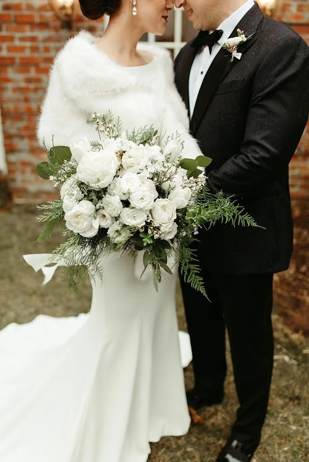 Closeup of a winter wedding bouquet with white flowers and greenery held by the bride and groom.