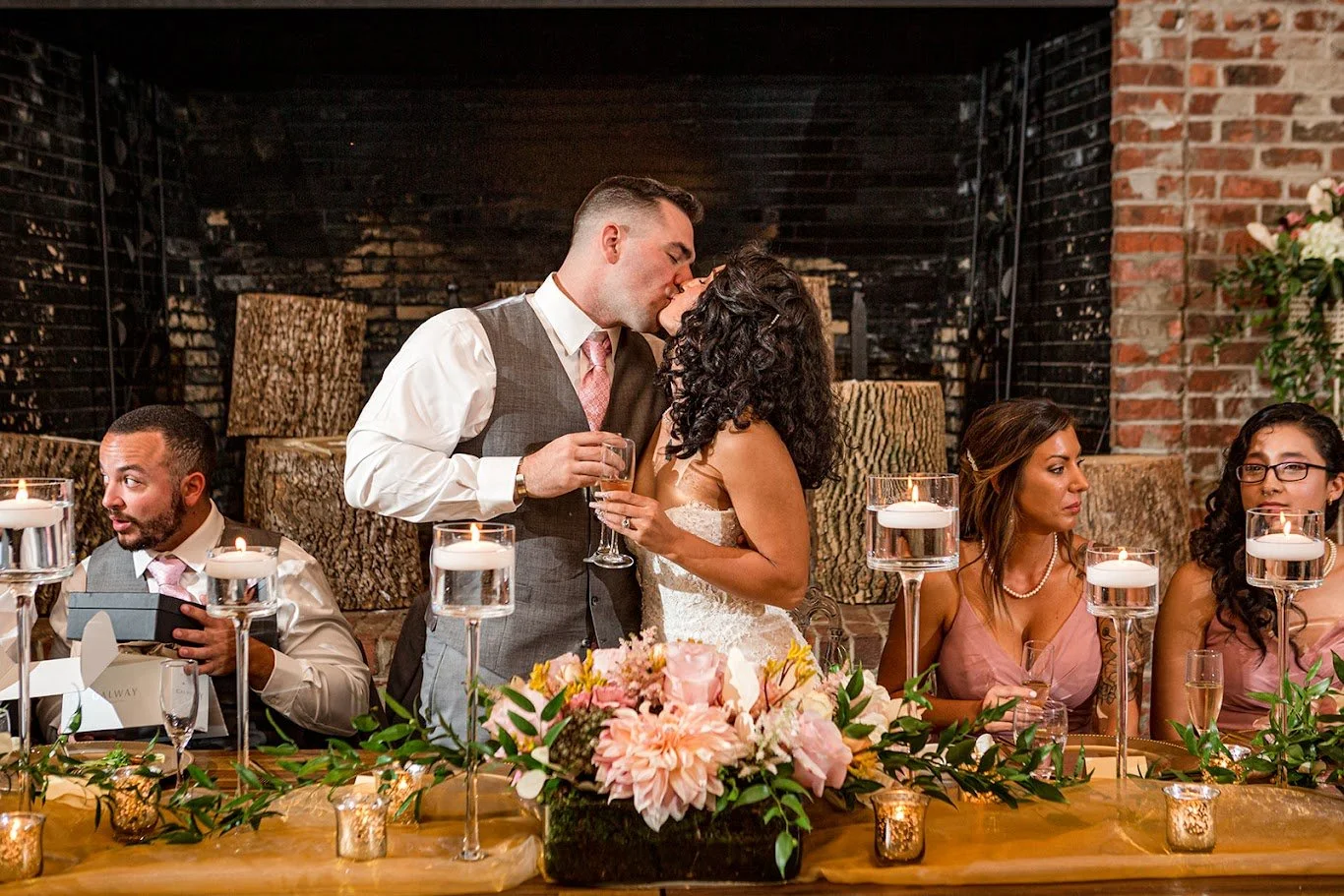 Bride and groom kissing at the sweetheart table in front of a brick fireplace with candlelight