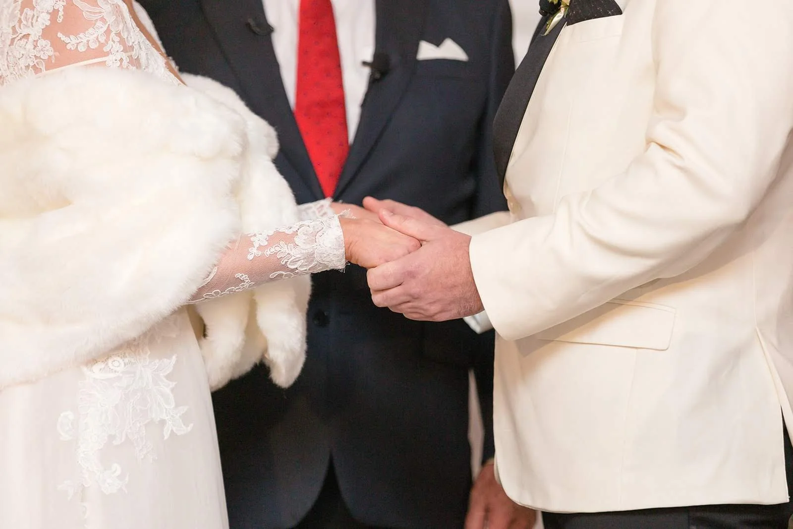 Closeup of the couple holding hands during the ceremony, bride wearing lace sleeves and fur wrap.