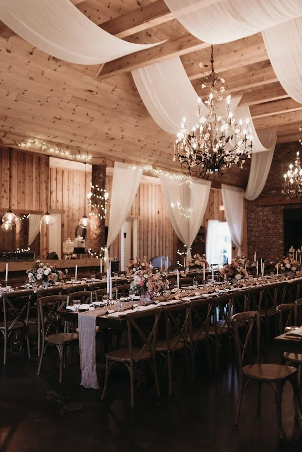 Long banquet tables set for a barn wedding reception under chandeliers and white fabric draping