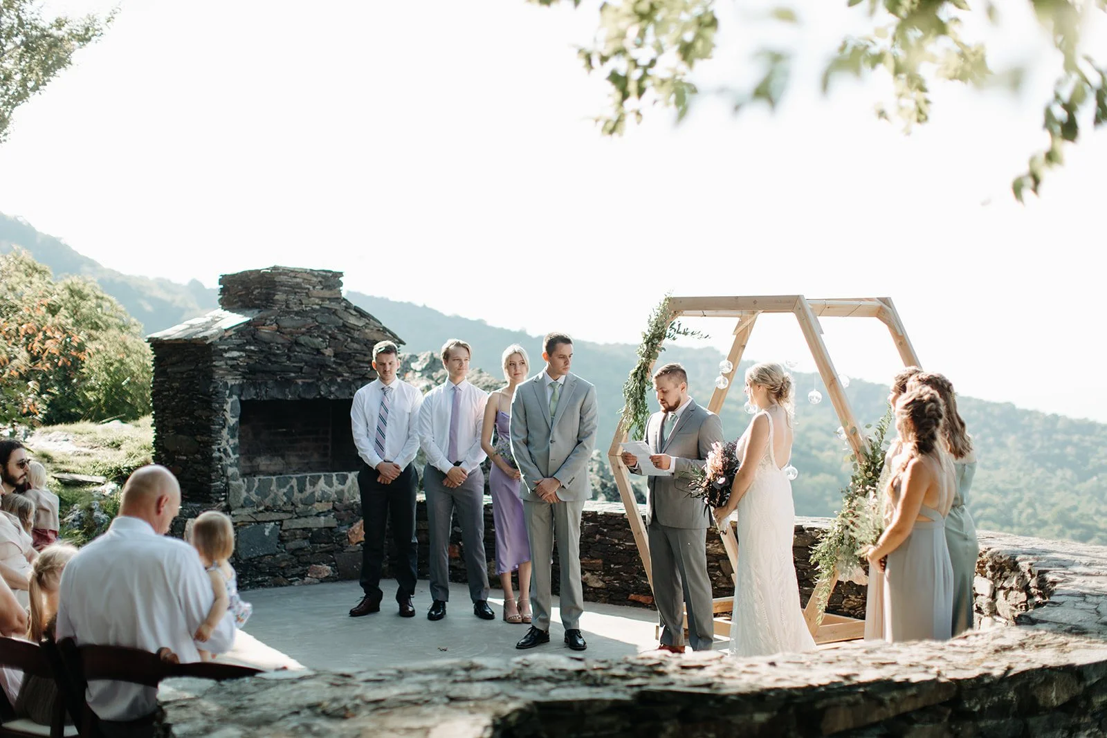 Groom reading vows to the bride during an outdoor mountain wedding ceremony beside a rustic stone fireplace.