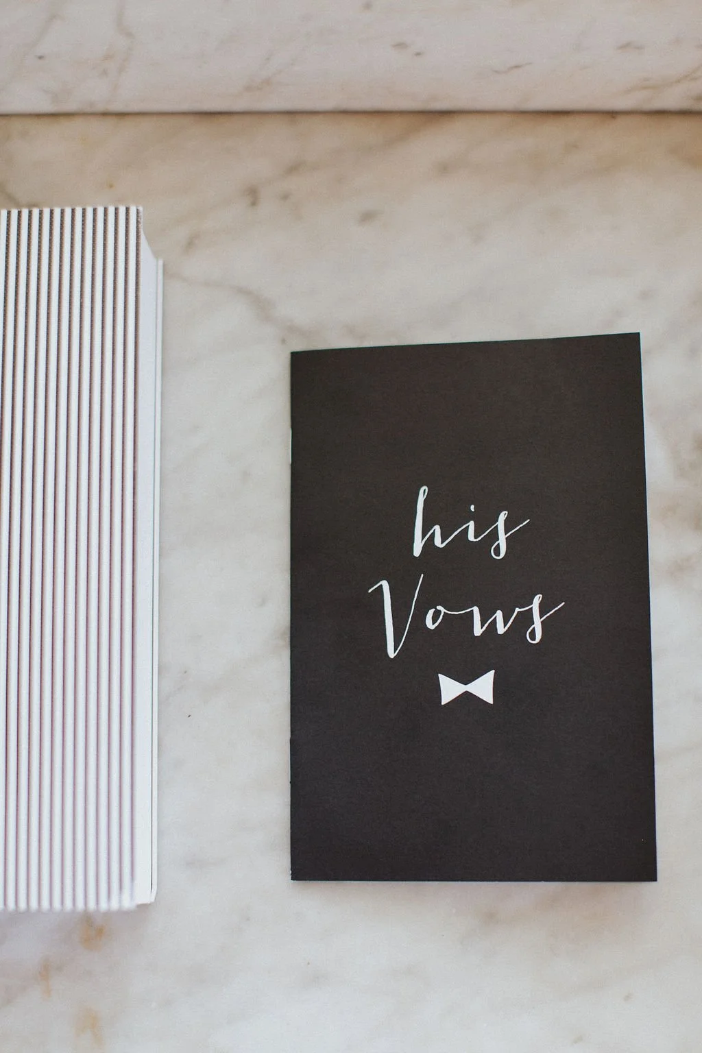 Black booklet reading “his vows” styled on a marble surface.