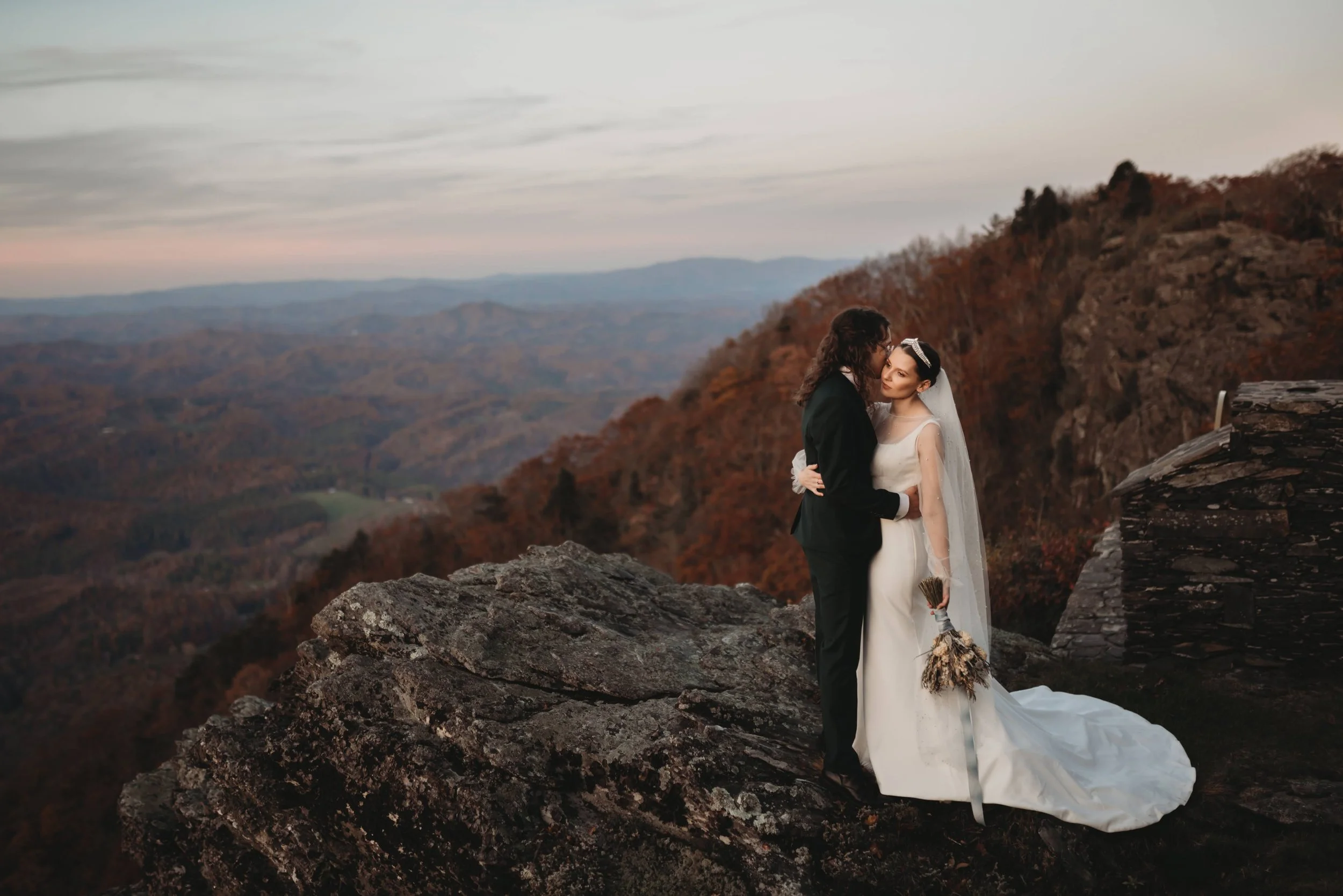 Bride and groom kissing on a rocky mountain cliff with sweeping fall-colored valley views