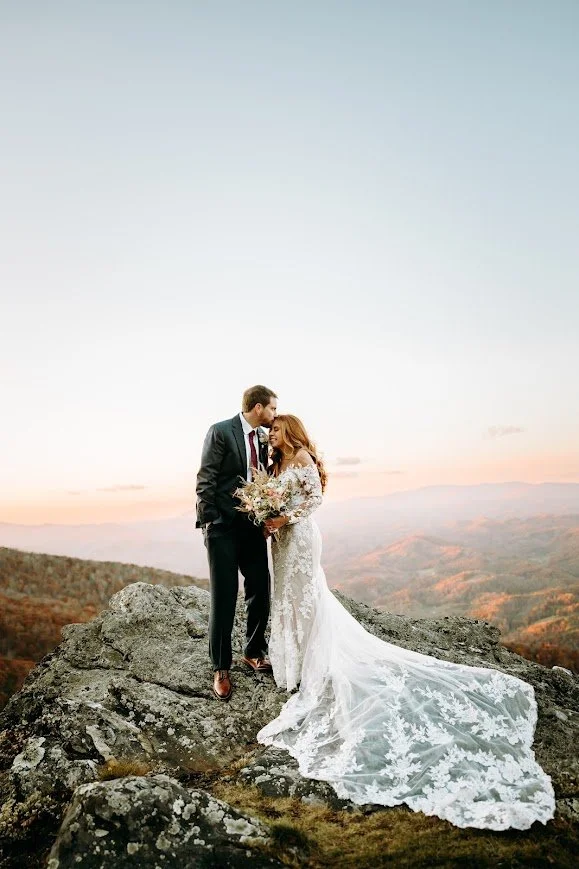 Bride and groom standing close on an overlook with the bride’s veil and train flowing