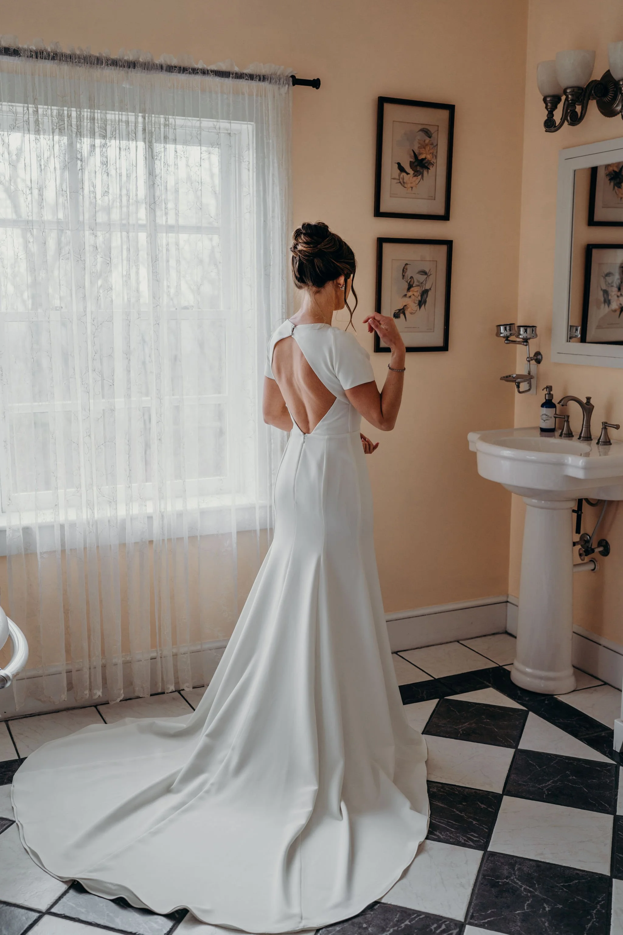 Bride standing in a bridal suite near a window, showing an open-back wedding dress and long train.