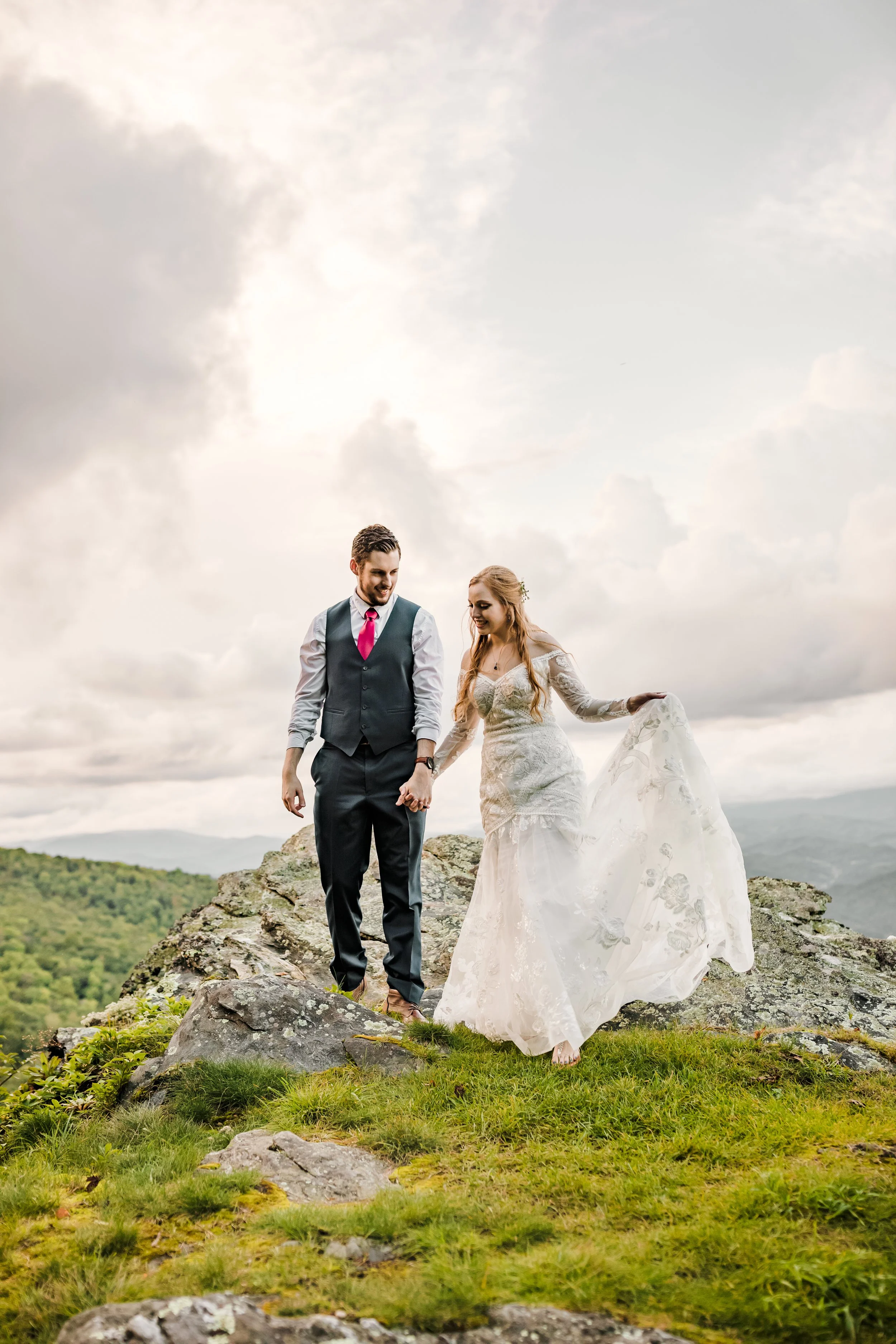 A smiling bride in a white lace gown and a groom in a vest and red tie hold hands, walking on a rocky mountain peak under a cloudy sky.