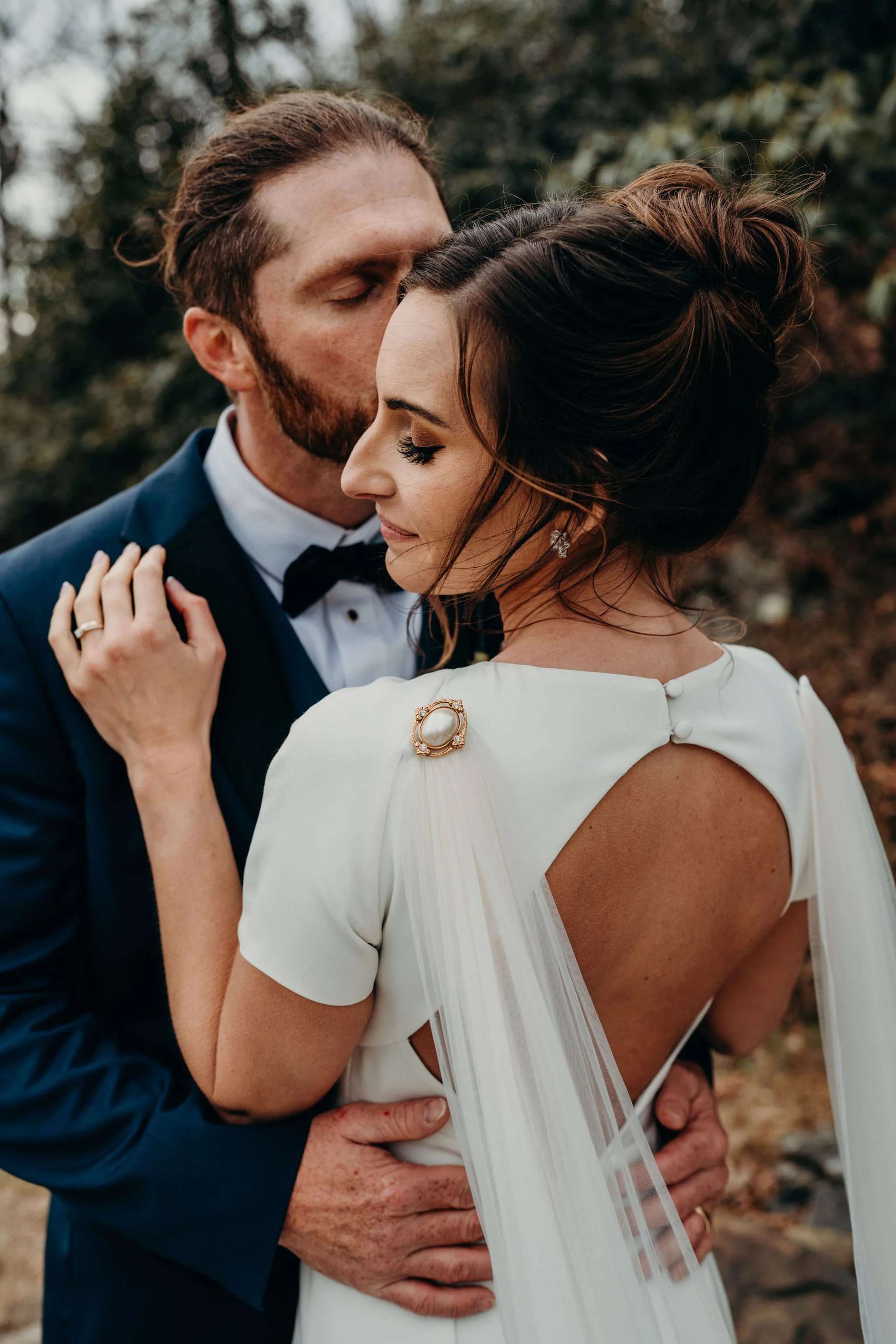 Closeup of the couple embracing, showing the bride’s veil fastened with a brooch and open-back dress.
