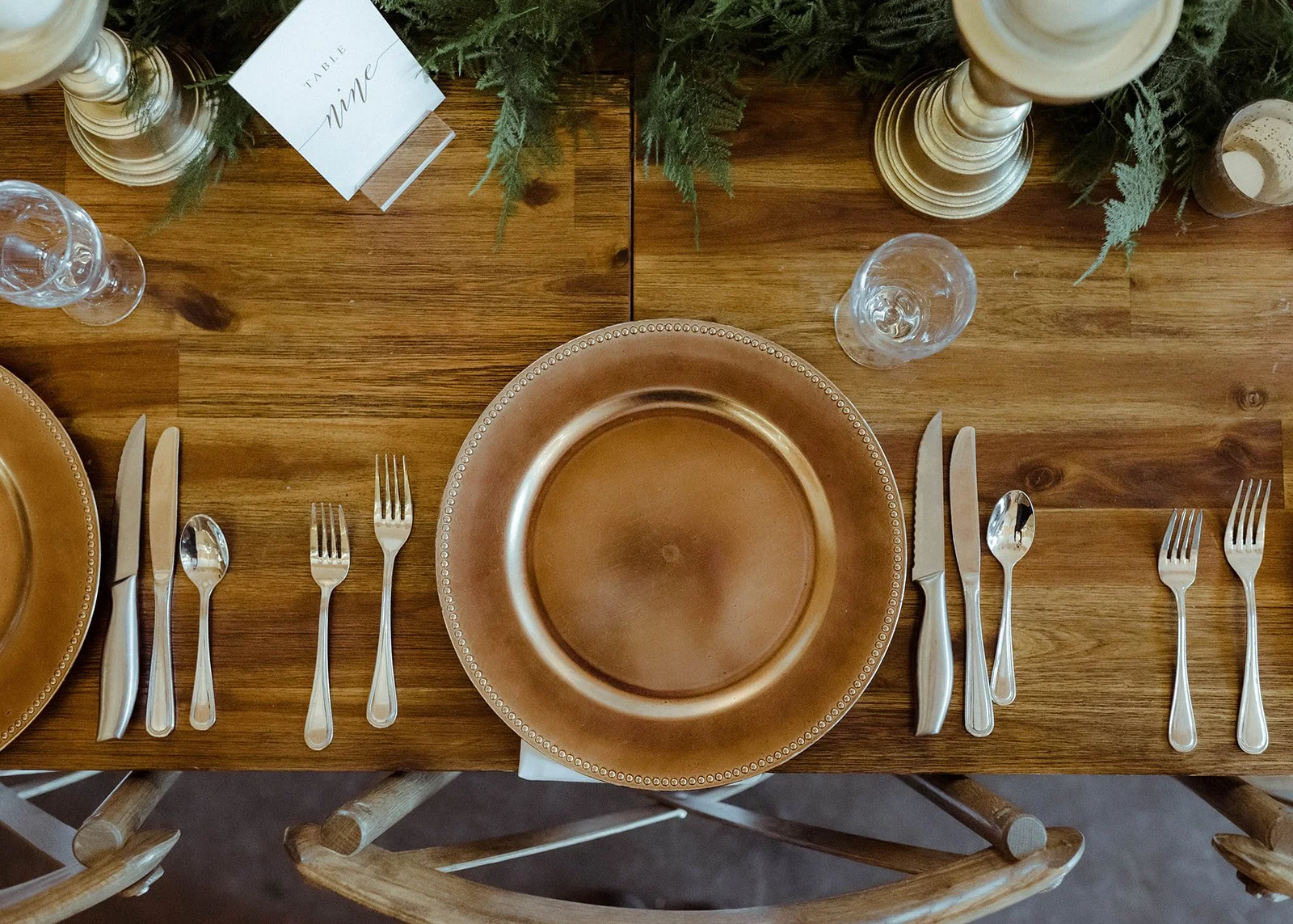 Neutral wedding reception place setting with brown plate, silver flatware, glassware, and greenery centerpiece.