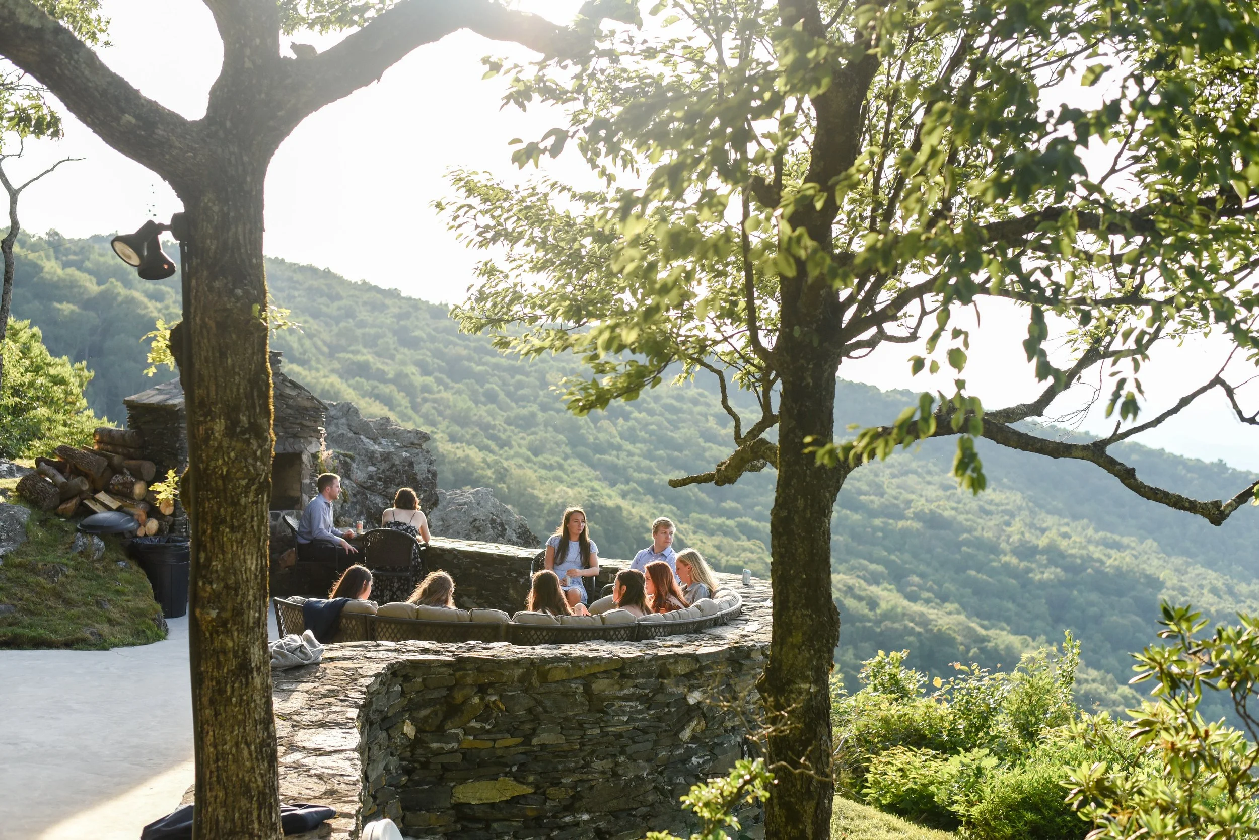 Corporate retreat guests relaxing in an outdoor mountain lounge area with stone walls, trees, and scenic valley views.