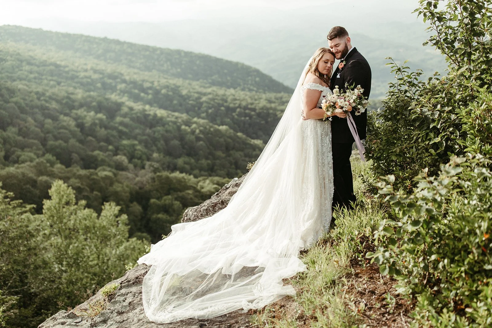 Bride and groom embracing on a mountain cliff with the bride’s long veil flowing and a bouquet in hand.