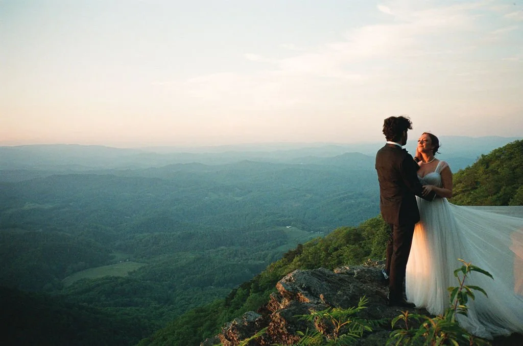 Bride and groom embracing on a Blue Ridge mountain cliff at sunset with layered valley views behind them.