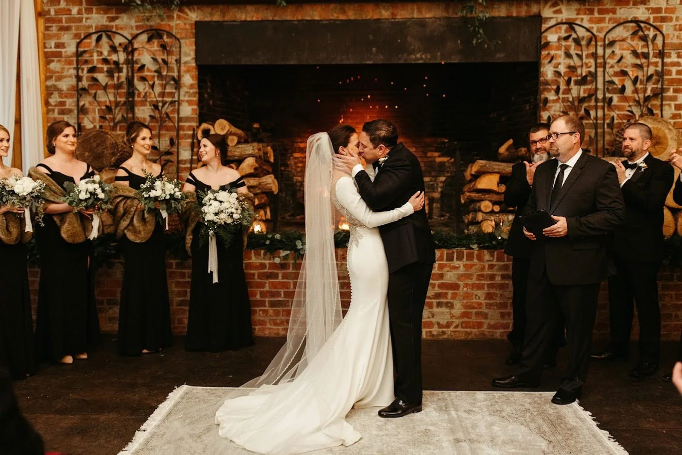 Bride and groom kissing in front of a fireplace during an indoor ceremony, with the bride’s long veil and train.