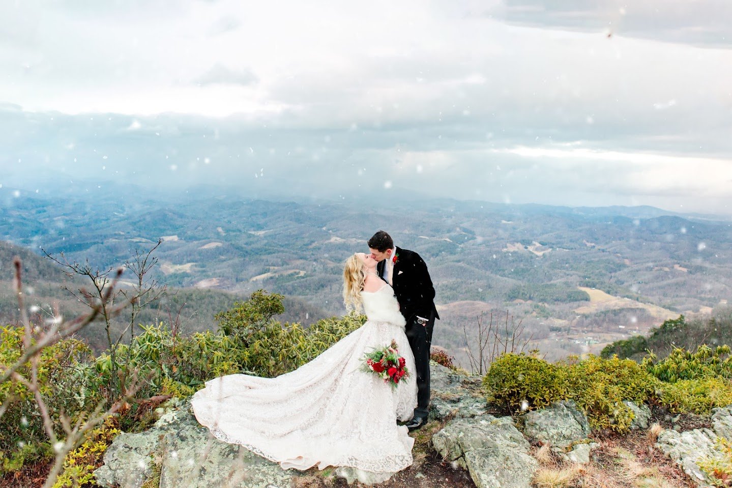 Bride and groom kissing on a mountaintop overlook with light snow falling and valley views.
