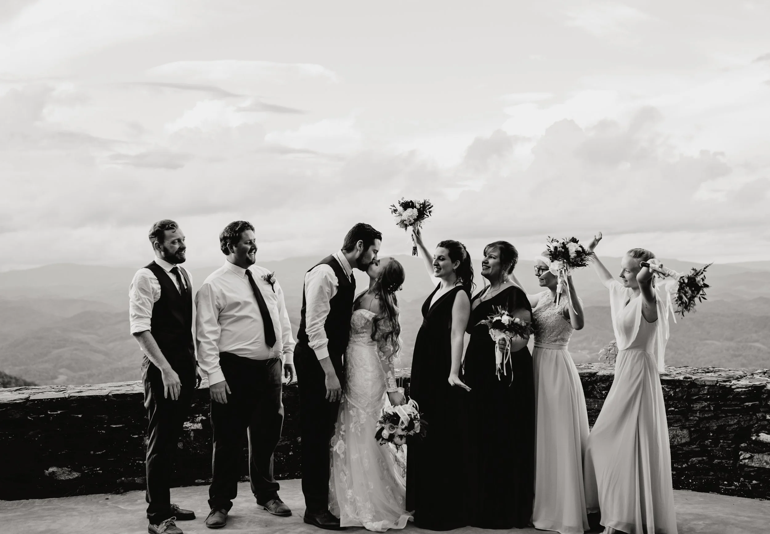Black and white: A wedding party celebrates outdoors. Bride and groom kiss, surrounded by groomsmen and bridesmaids with bouquets, mountains in background.