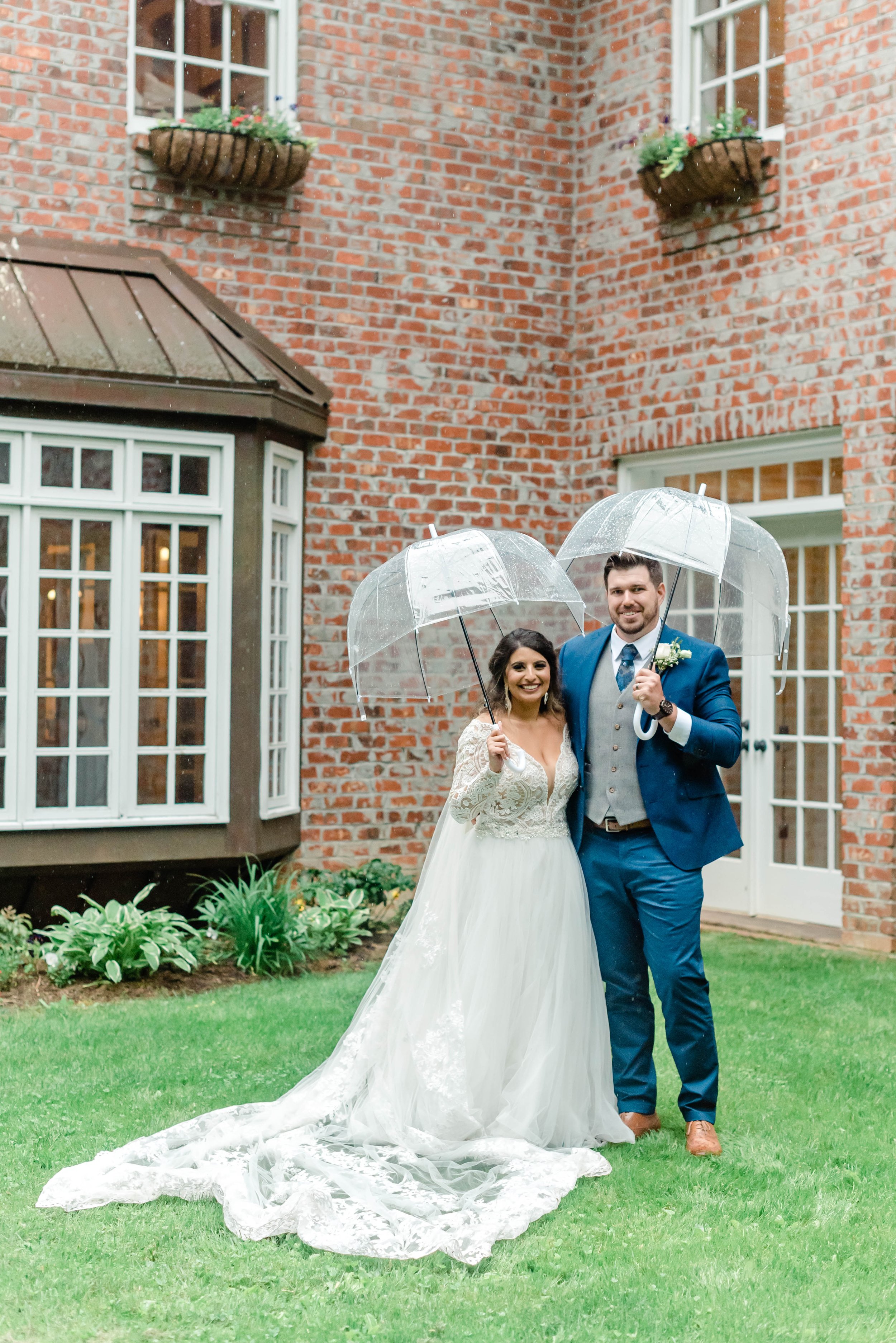 Bride and groom standing under clear umbrellas outside a brick wedding venue after the ceremony.