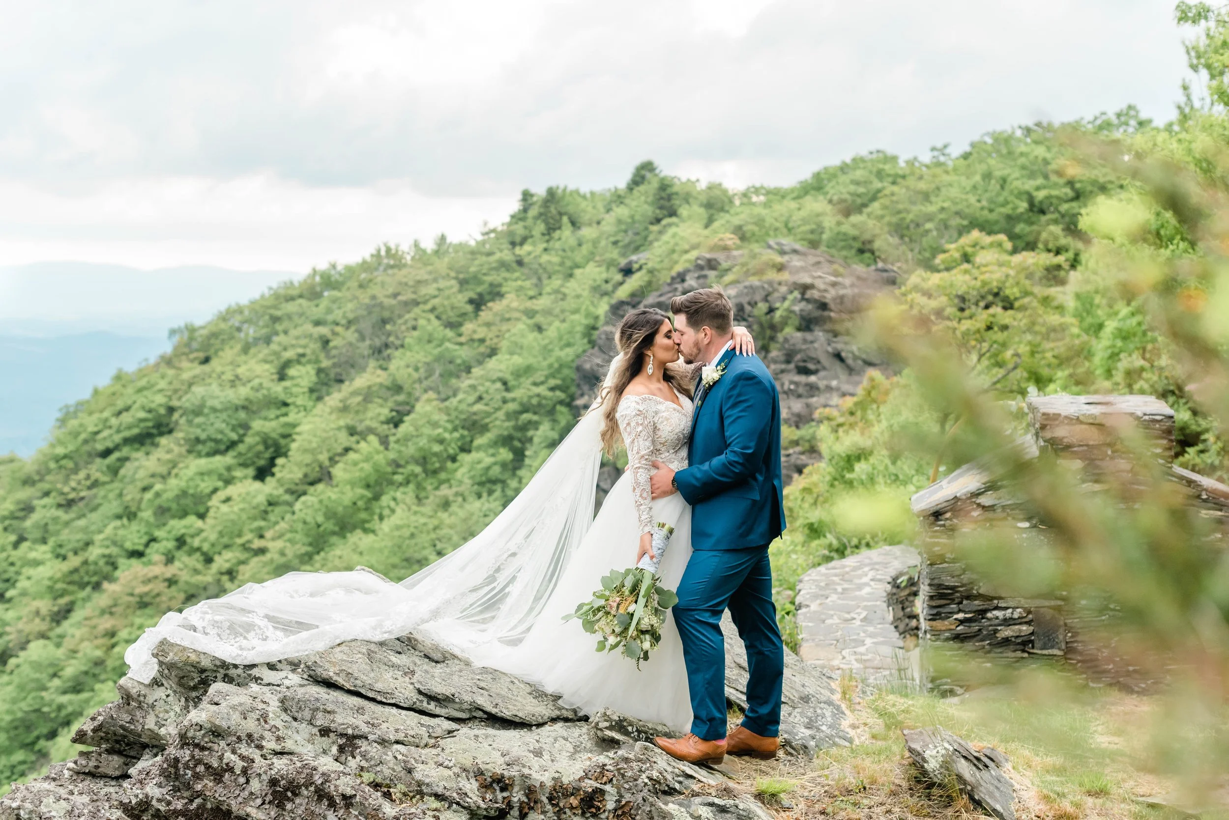 Bride and groom kiss on a rocky overlook, her long veil trailing, with green mountains and cloudy sky behind.