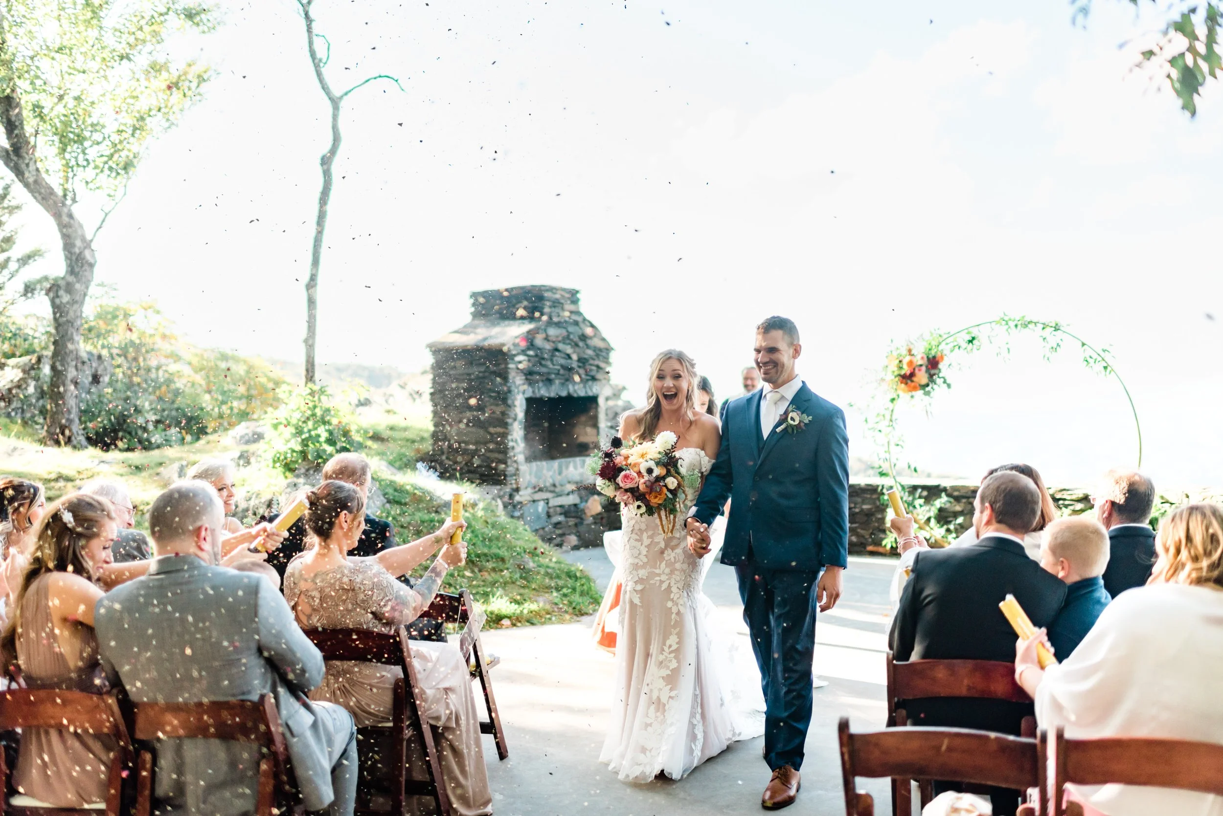 Bride and groom walking down the aisle during their ceremony exit as guests celebrate with confetti outdoors.