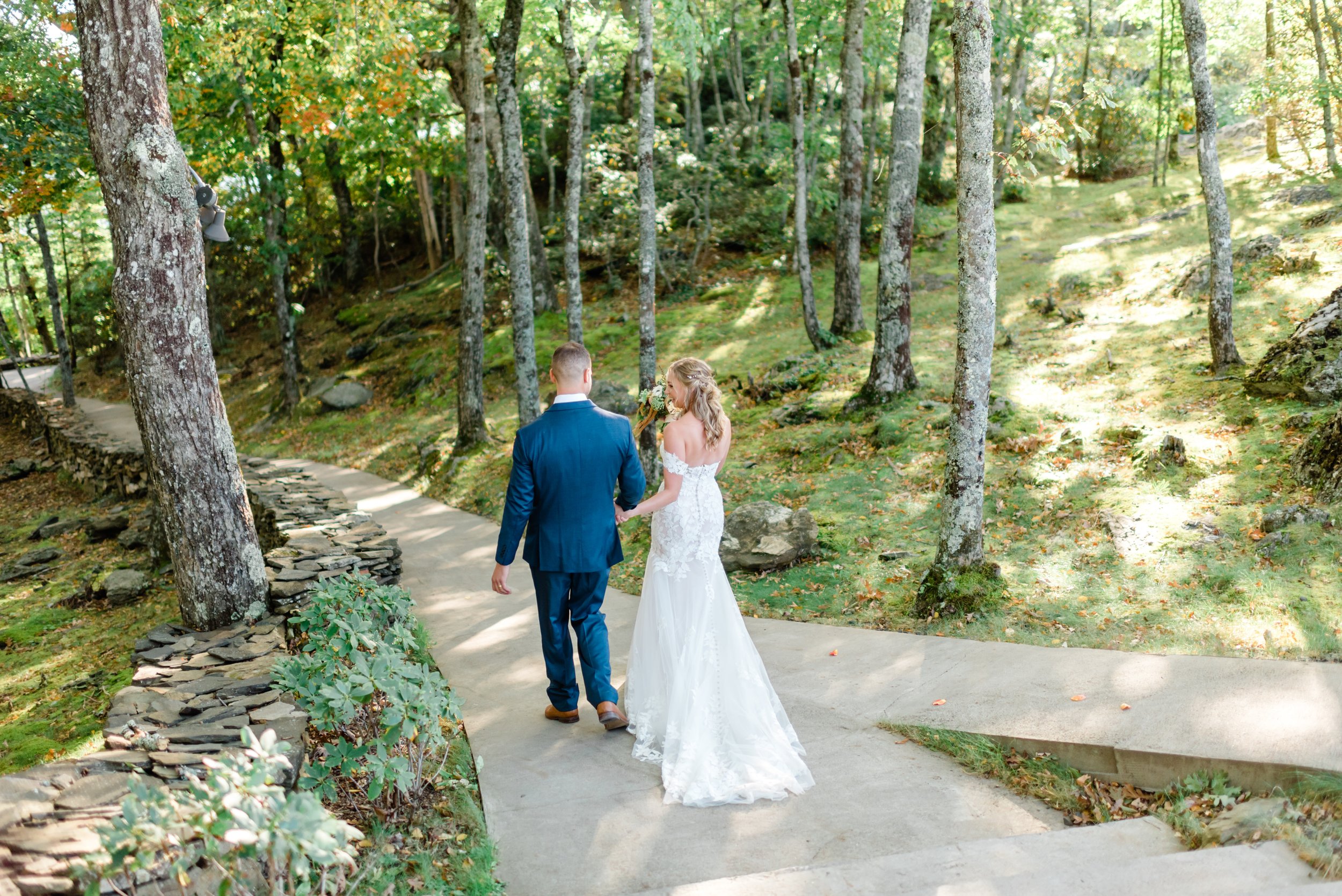 Bride and groom walking hand in hand through a wooded mountain trail with stone walls, trees, and soft sunlight.