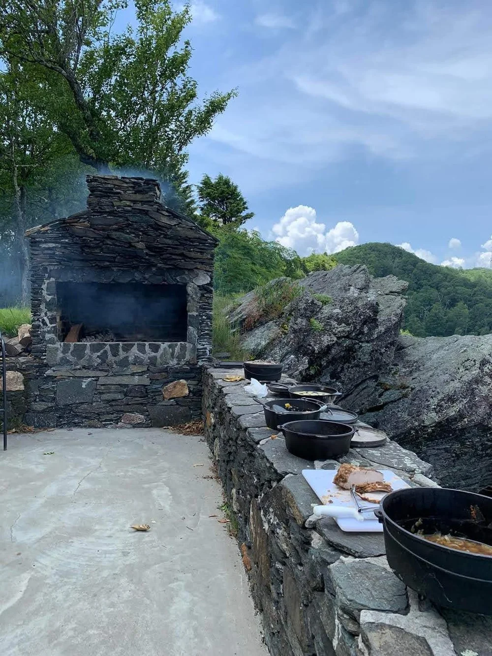 Outdoor cooking setup in the North Carolina mountains with a stone fireplace/oven and several cast iron pots on a stone wall overlooking a scenic landscape.