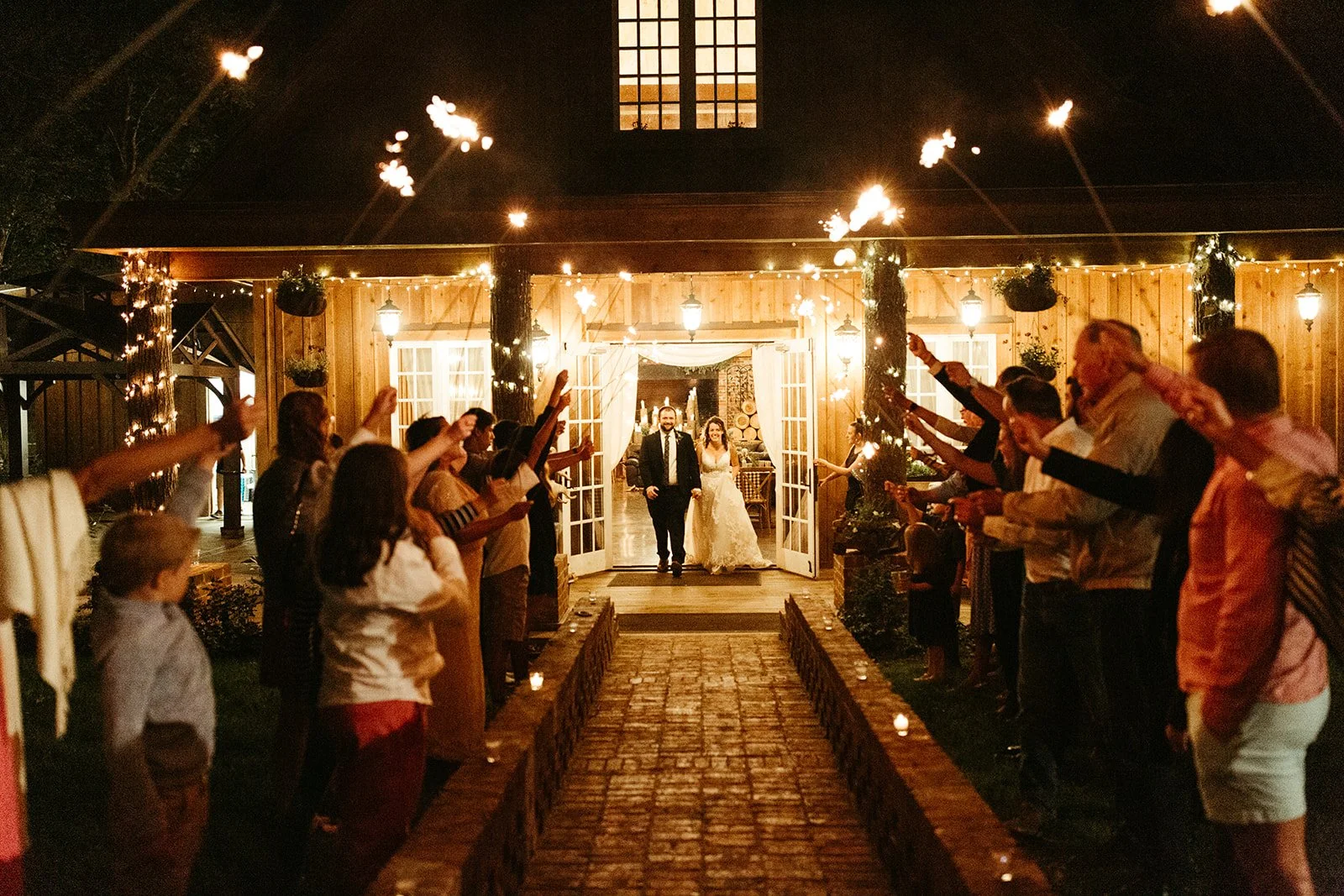 Newlyweds walking through a sparkler exit outside a rustic barn wedding reception at night.