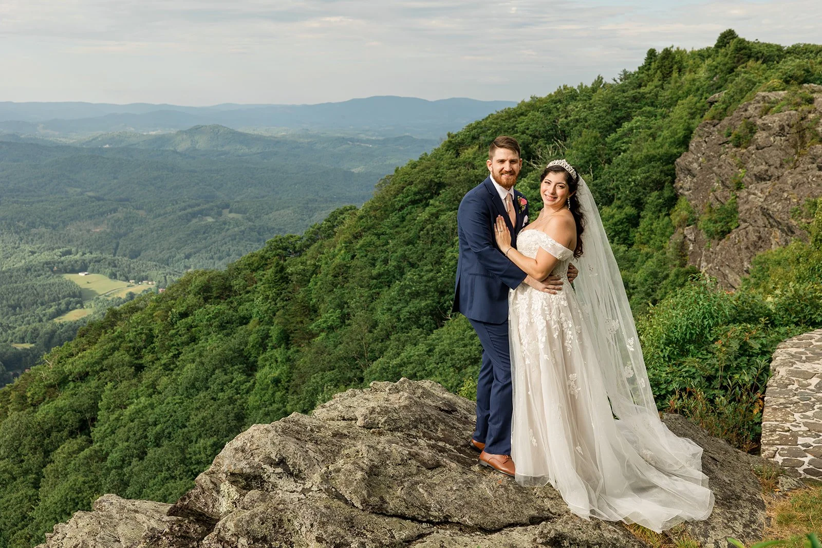 Bride and groom posing together on a rocky mountain overlook with sweeping green valley views behind them.Bride and groom posing together on a rocky mountain overlook with sweeping green valley views behind them.