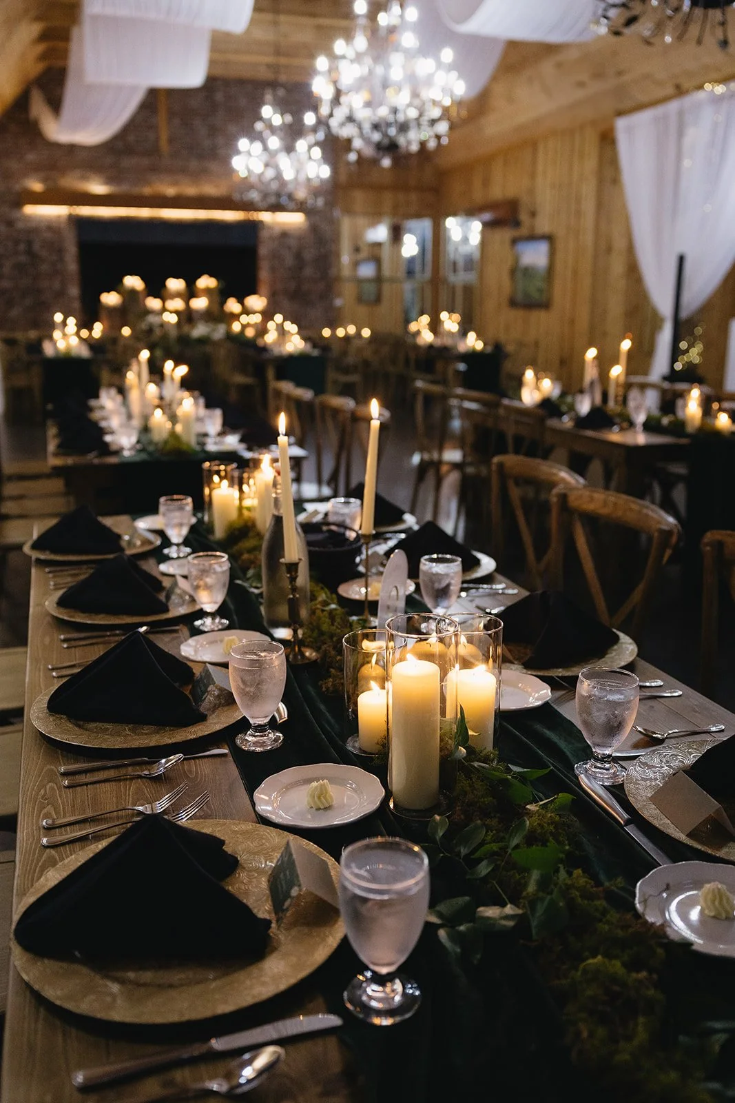 Candlelit wedding reception table with black napkins, glassware, taper candles, and greenery on a wooden table