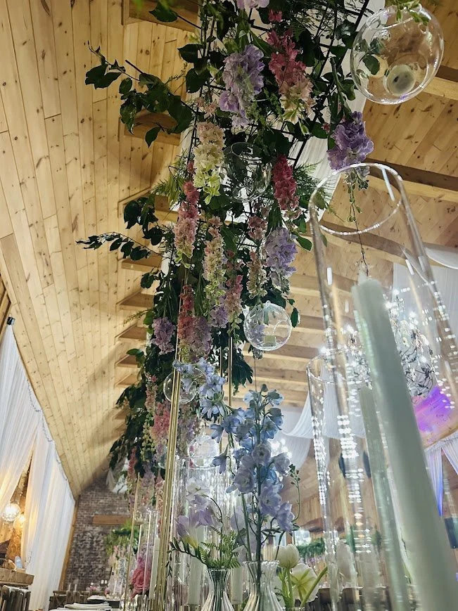 Dramatic hanging floral installation with greenery and purple blooms suspended from a wooden ceiling above reception tables.
