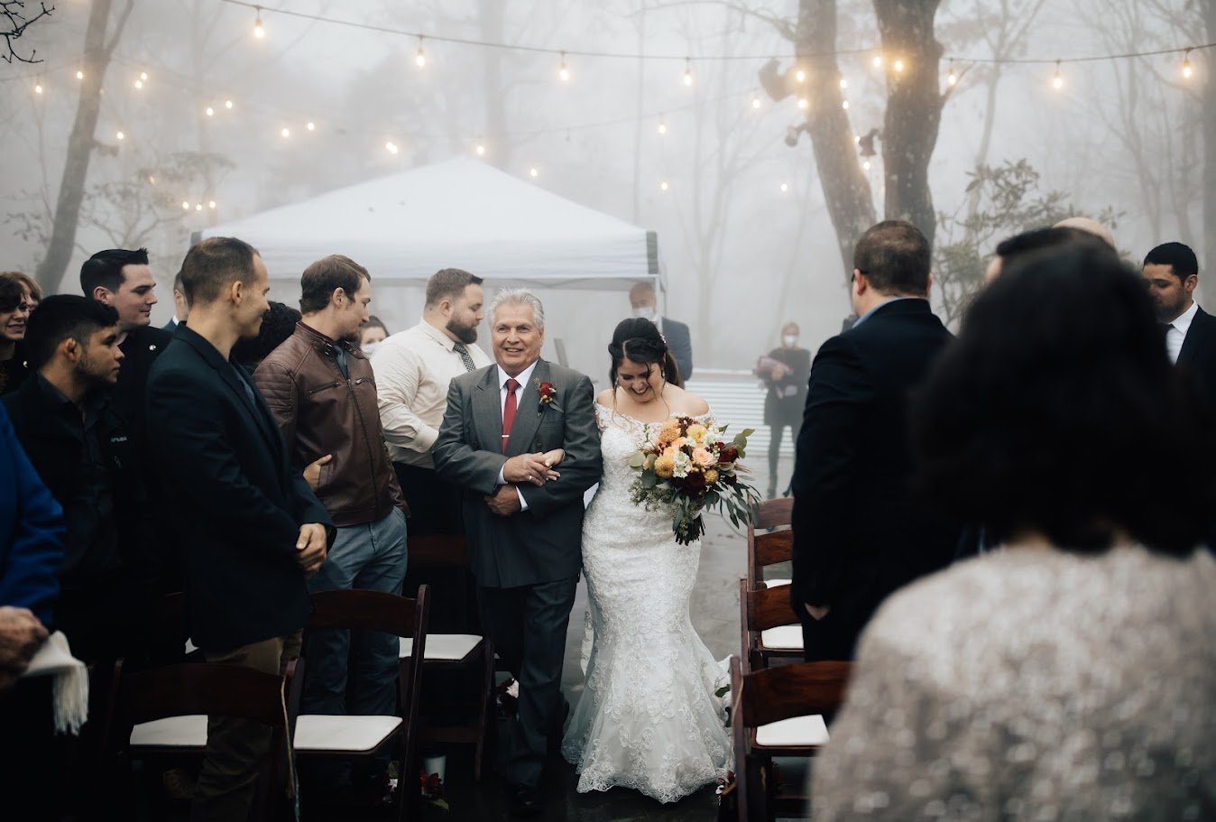 Bride walking down the aisle with her father in a foggy outdoor ceremony with string lights.