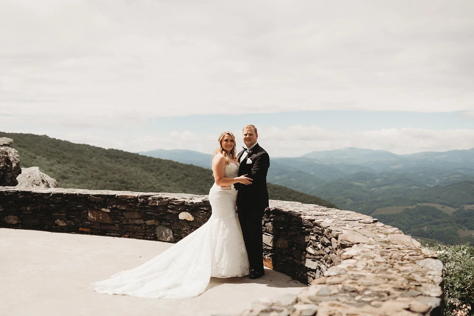 Bride and groom embrace by a stone wall with expansive mountain views in the background.