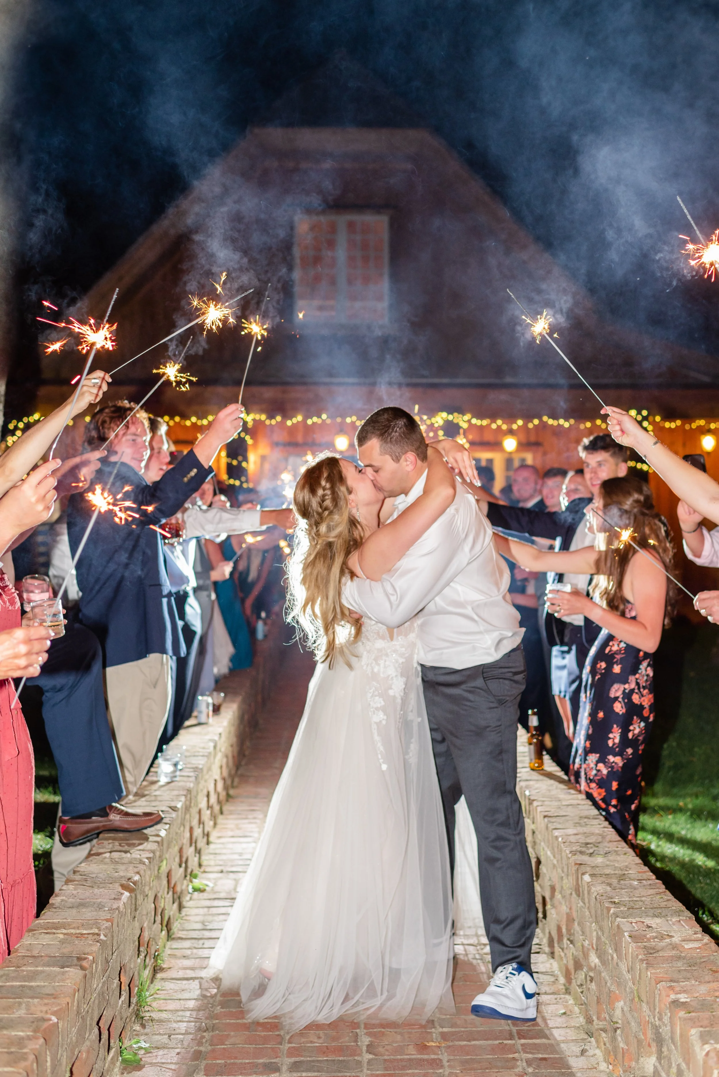Bride and groom kissing during a nighttime sparkler exit as wedding guests celebrate along a stone walkway.