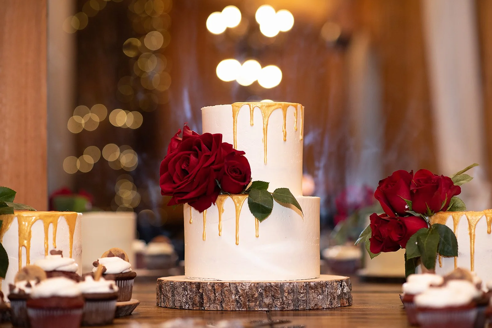 Two-tier white wedding cake with gold drip icing and red roses displayed on a wooden slice stand
