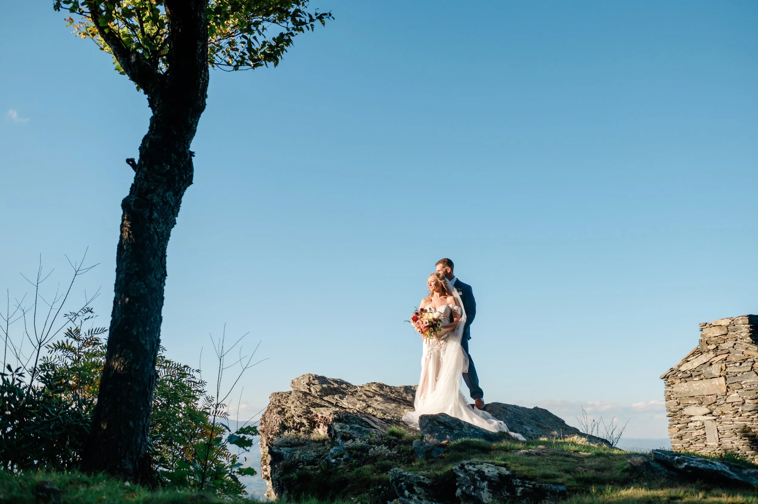Bride and groom standing together on a rocky mountain overlook with blue sky, trees, and stone ruins nearby.