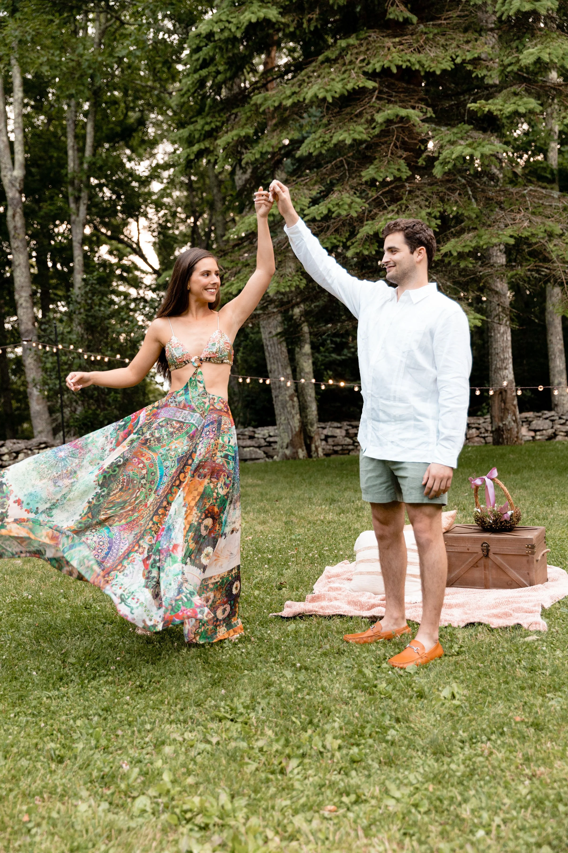 Couple twirling together on a lawn during a casual welcome party with string lights and trees behind them.